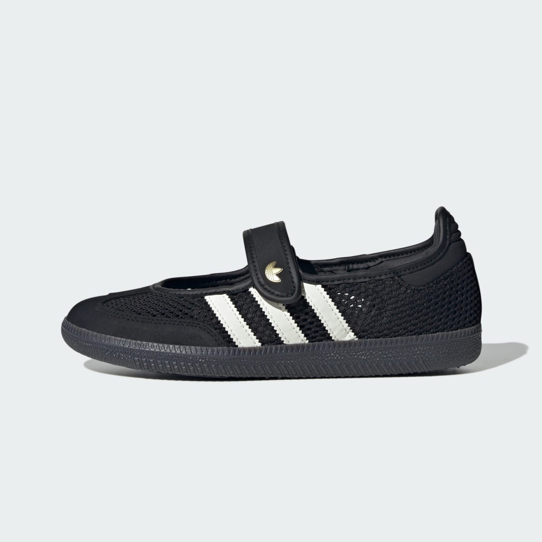 A black knit slip-on adidas samba jane shoe with three white stripes and a hook-and-loop strap featuring a gold adidas logo, shown from the side on a light gray background.