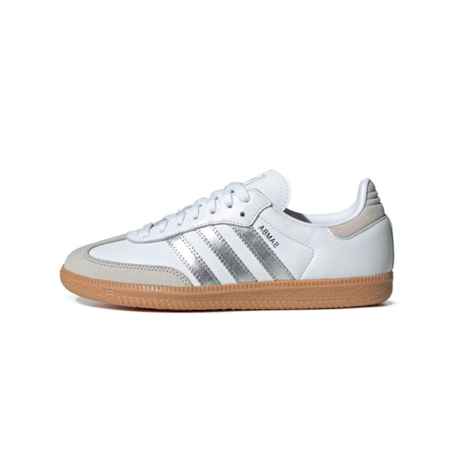 A white Adidas Samba sneaker with metallic silver stripes, a white leather upper, and a classic brown gum sole, shown in profile against a white background.