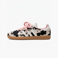 Adidas samba cow print sneaker shown in profile against a plain white background. The shoe features a black and white cow-print pattern on the upper, white side stripes, fuzzy light pink laces, a pink collar lining, and a brown gum sole.