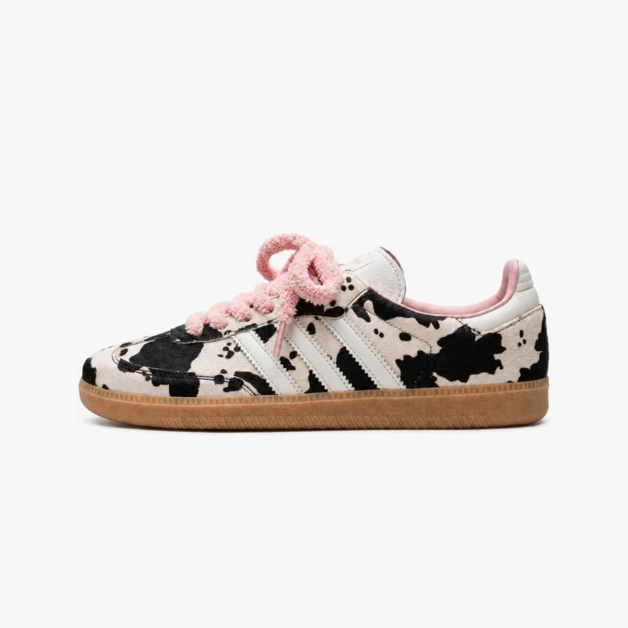 Adidas samba cow print sneaker shown in profile against a plain white background. The shoe features a black and white cow-print pattern on the upper, white side stripes, fuzzy light pink laces, a pink collar lining, and a brown gum sole.