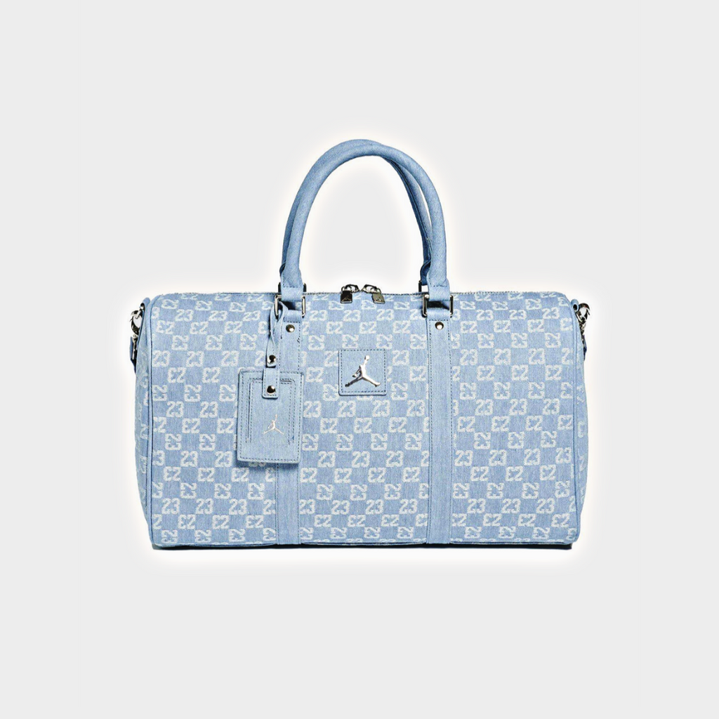 A light blue monogram duffel bag with silver hardware, featuring a top handle, a matching luggage tag, and a small silver Jordan logo on the front, set against a light gray background.