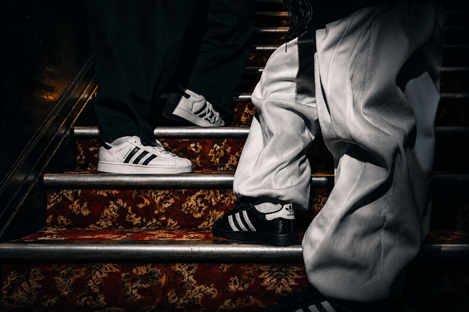 Two people standing on patterned indoor stairs, shown from the knees down. One person wears white Adidas Superstars with black stripes, and the other wears black Adidas Superstars with white stripes, paired with light gray sweatpants.