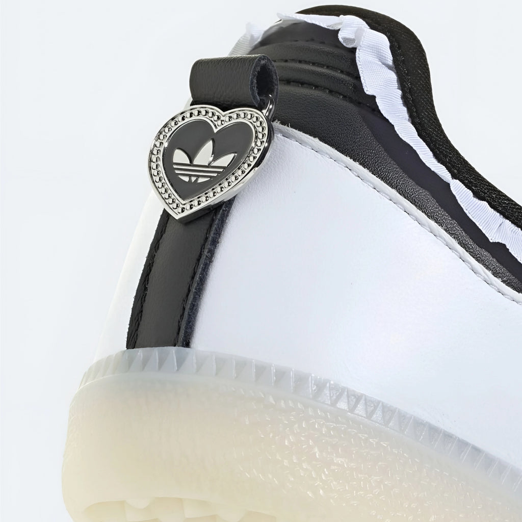 Close-up of a white and black Adidas sneaker featuring a heart-shaped silver metal charm with the Adidas Trefoil logo on the heel strap.
